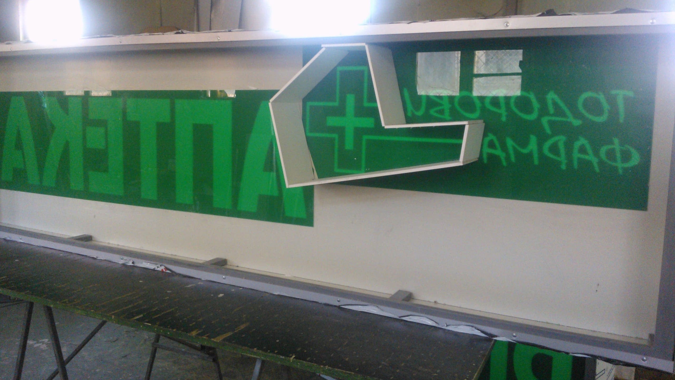 Вътрешна част на табела в процес на изработка, показваща зелен шаблон с огледални надписи и триизмерна бяла фигура на кръст.