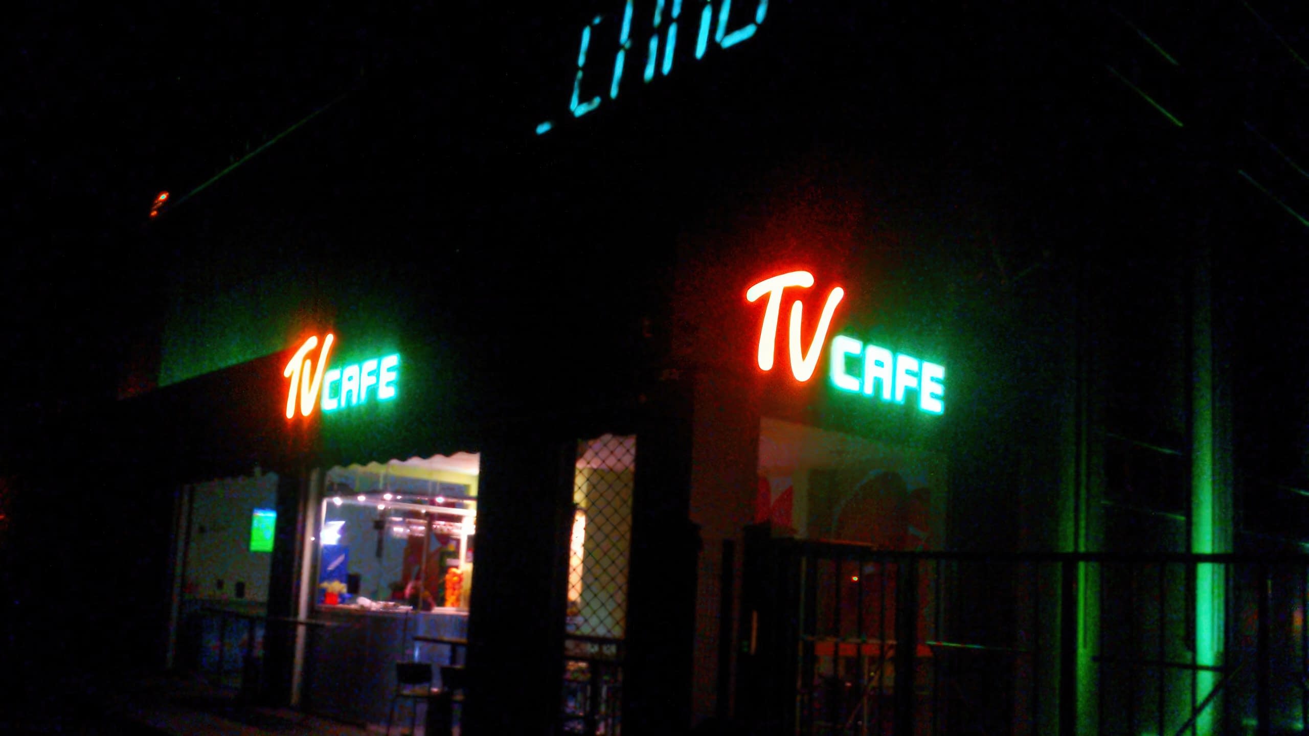 Светещи в оранжево и зелено обемни букви с надпис "TV CAFE" на фасадата на сграда през нощта.