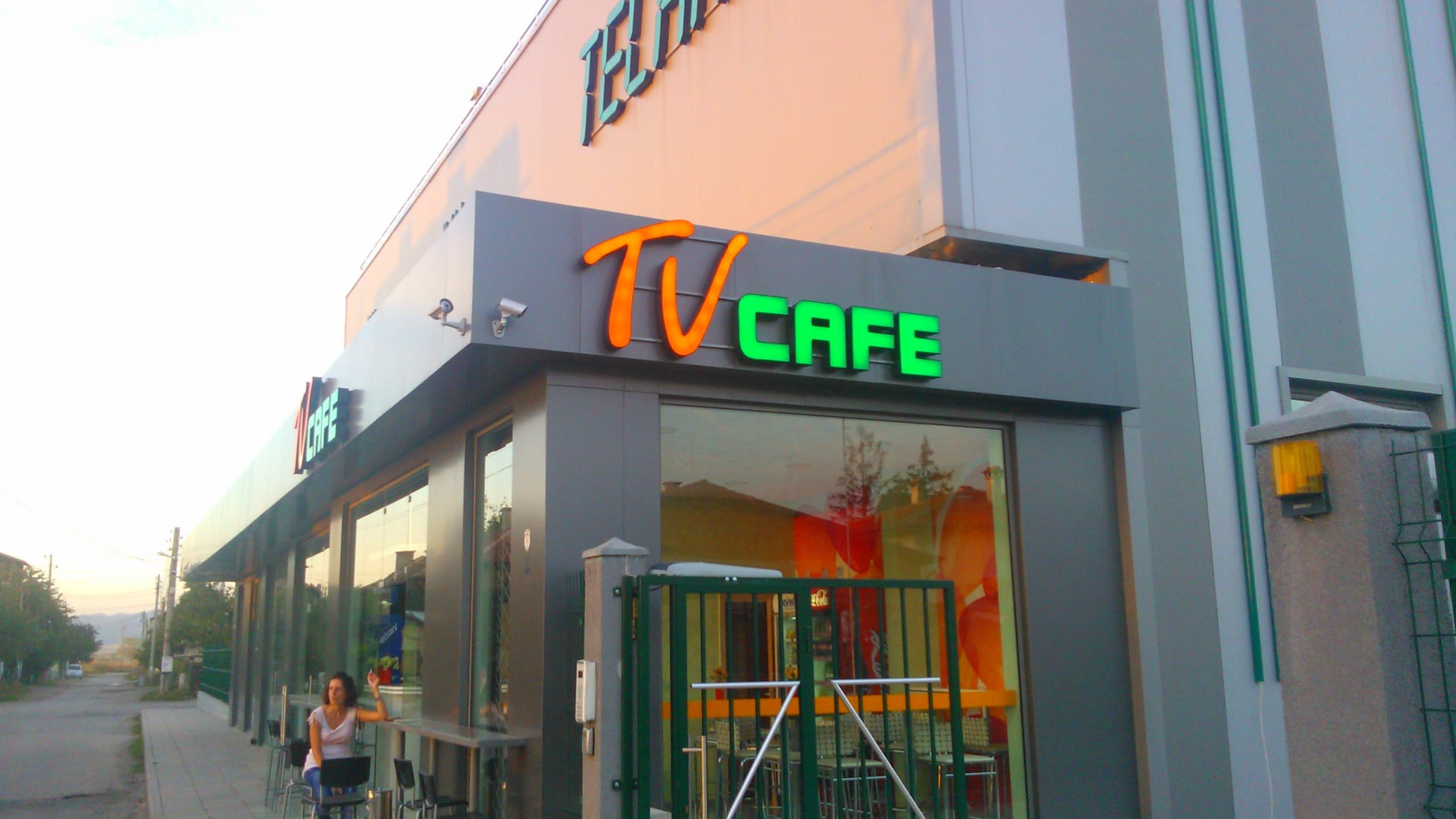 Външен изглед на сграда с надпис "TV CAFE" от обемни букви в оранжево и зелено, заснет през деня.
