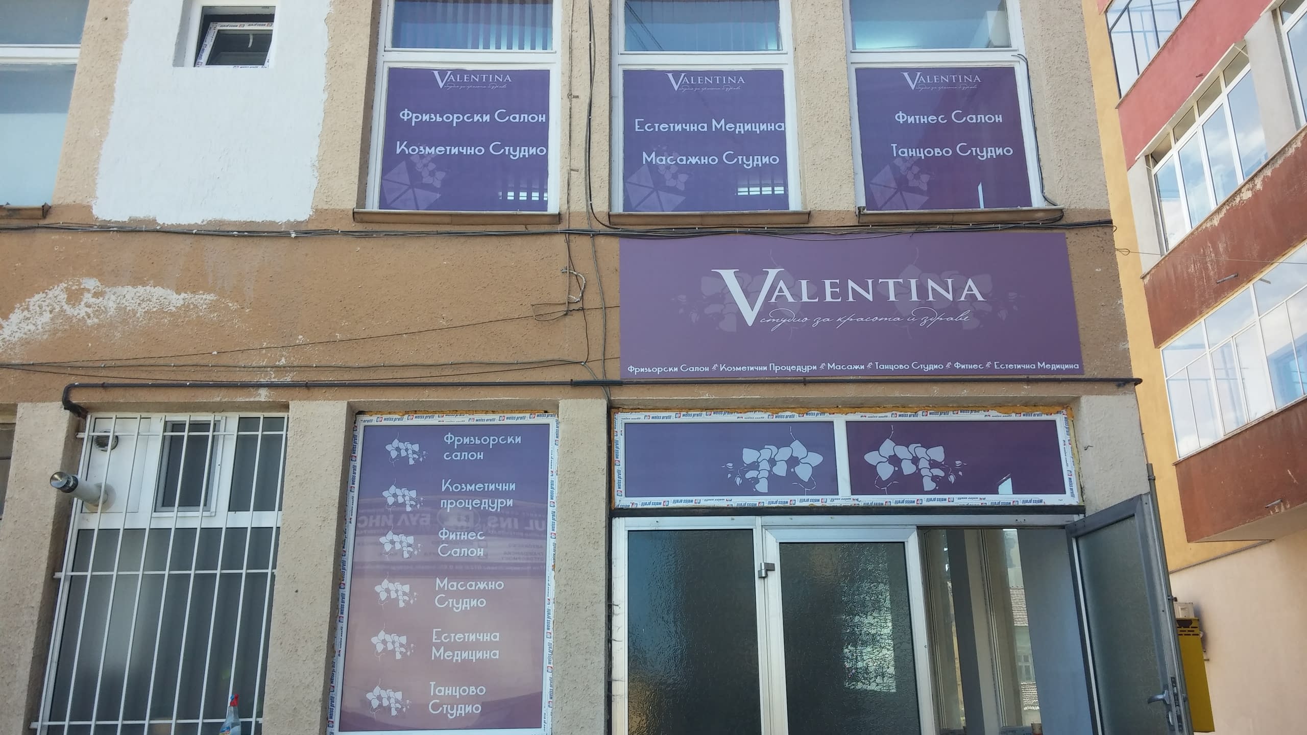 Фасада на сграда с лилава табела и стикери на прозорците, изброяващи услугите на студио за красота.