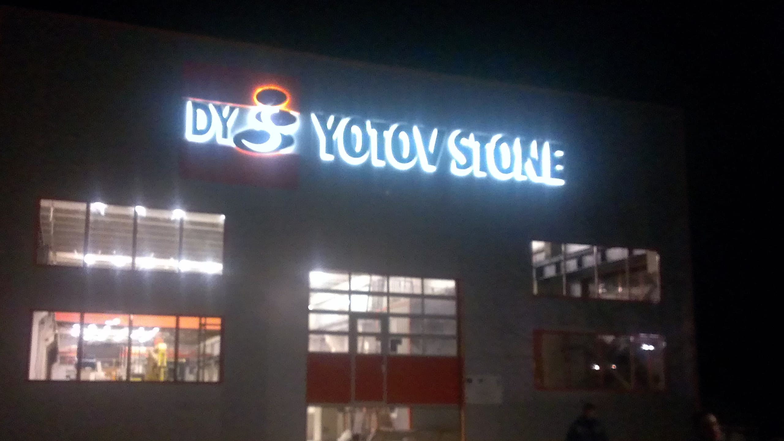 Ярко светещо лого и надпис "Yotov Stone" на фасадата на сграда в тъмната част на денонощието.