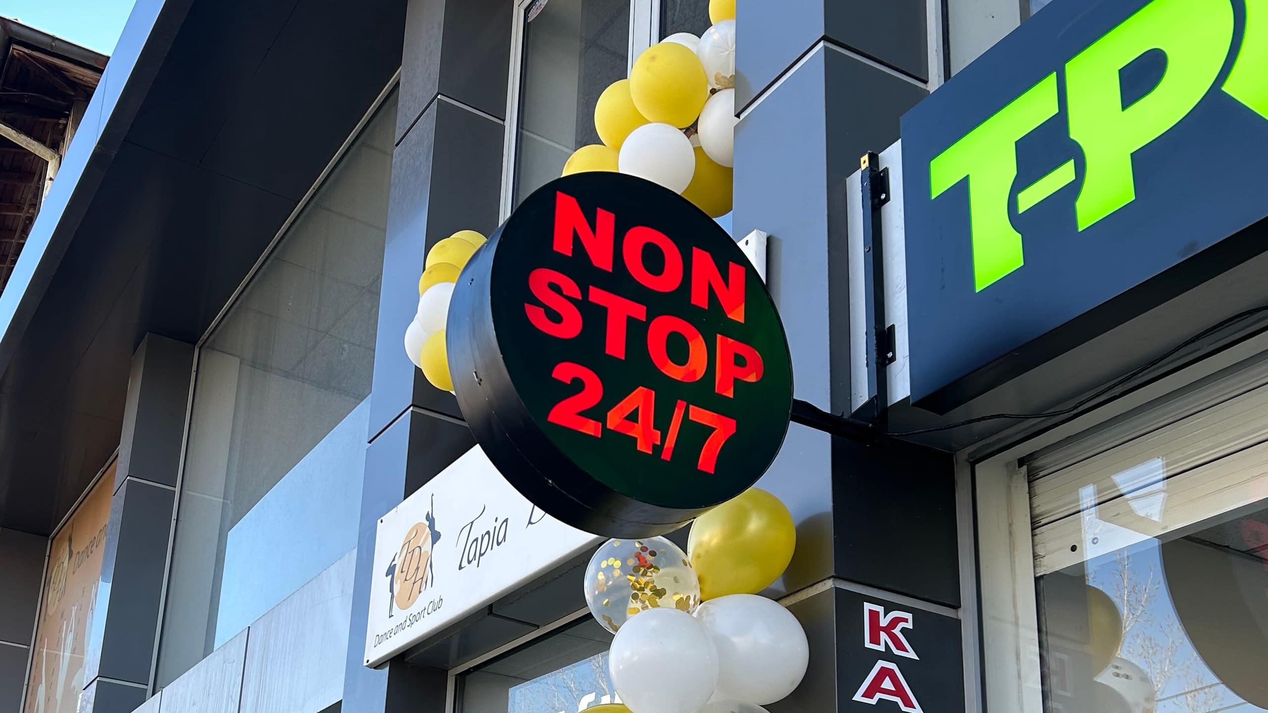 Двустранна кръгла светеща LED табела с надпис "NON STOP 24/7" пред денонощен магазин.