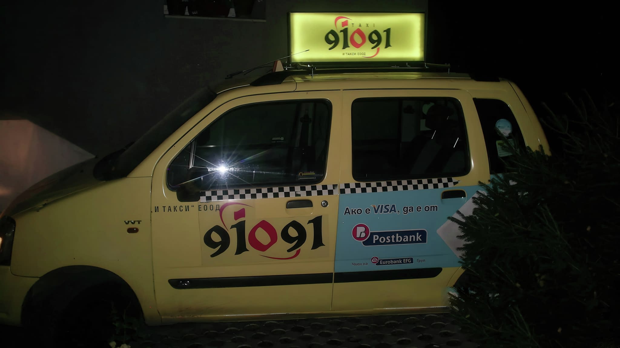 Светеща реклама върху жълто такси 91091 с брандиране от Postbank и реклама на Visa, заснето през нощта.