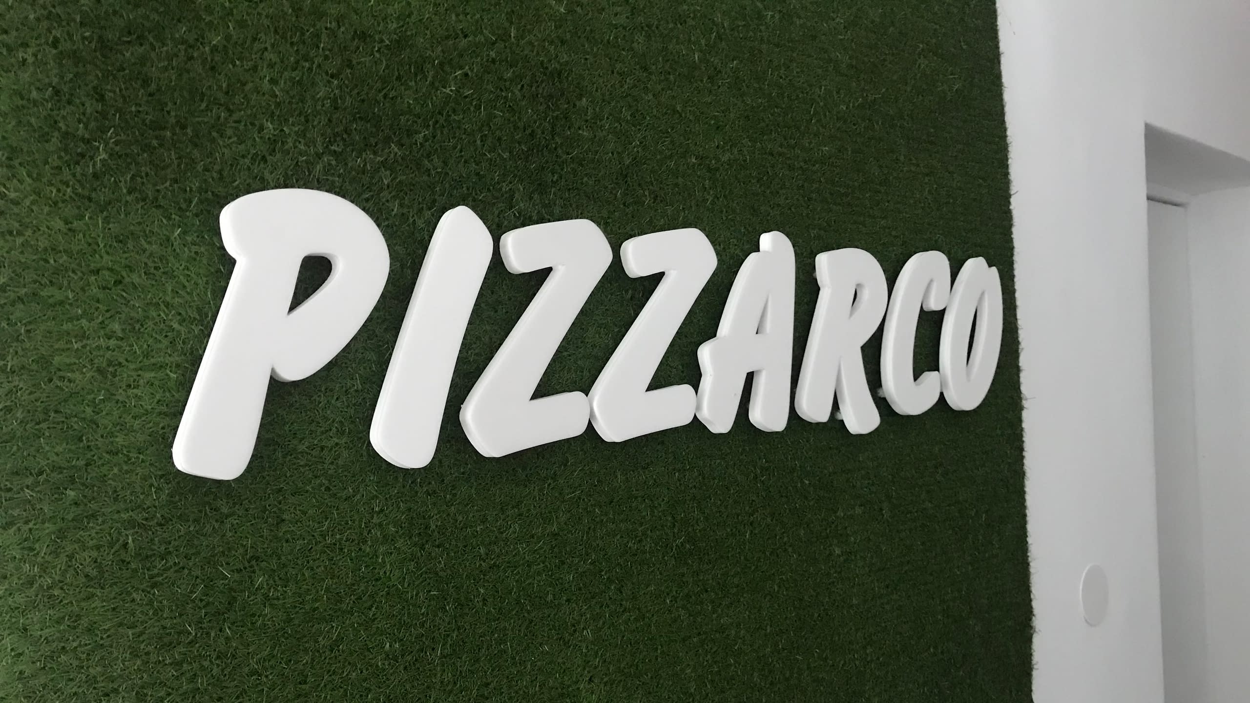 Бели обемни букви с надпис "PIZZARCO", монтирани върху стена с изкуствена трева. Ефектна интериорна реклама за заведение или офис.