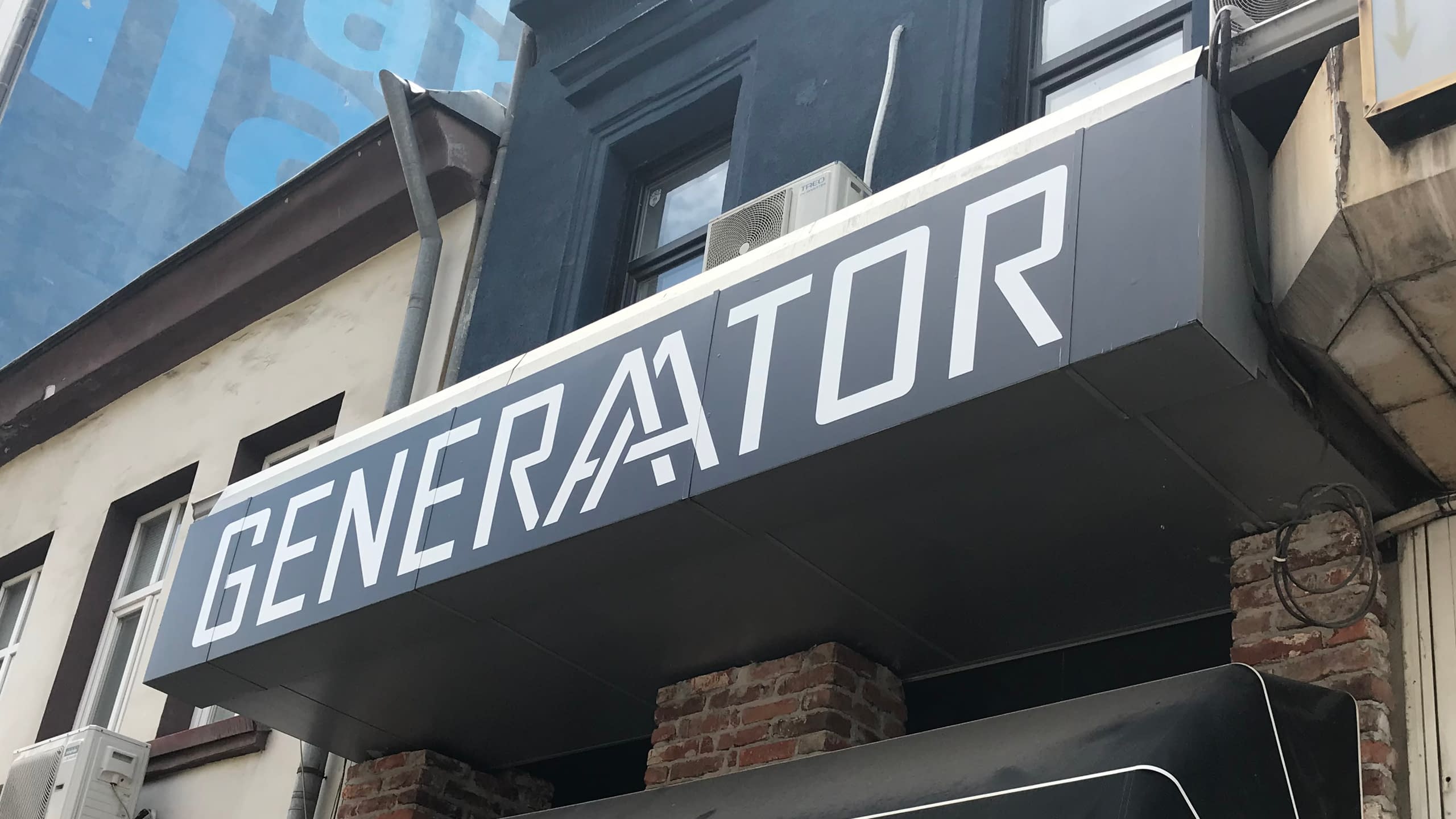 Модерен рекламен надпис "GENERATOR" с бели букви върху тъмна табела. Външна реклама за заведение или креативно пространство със стилен и изчистен дизайн.
