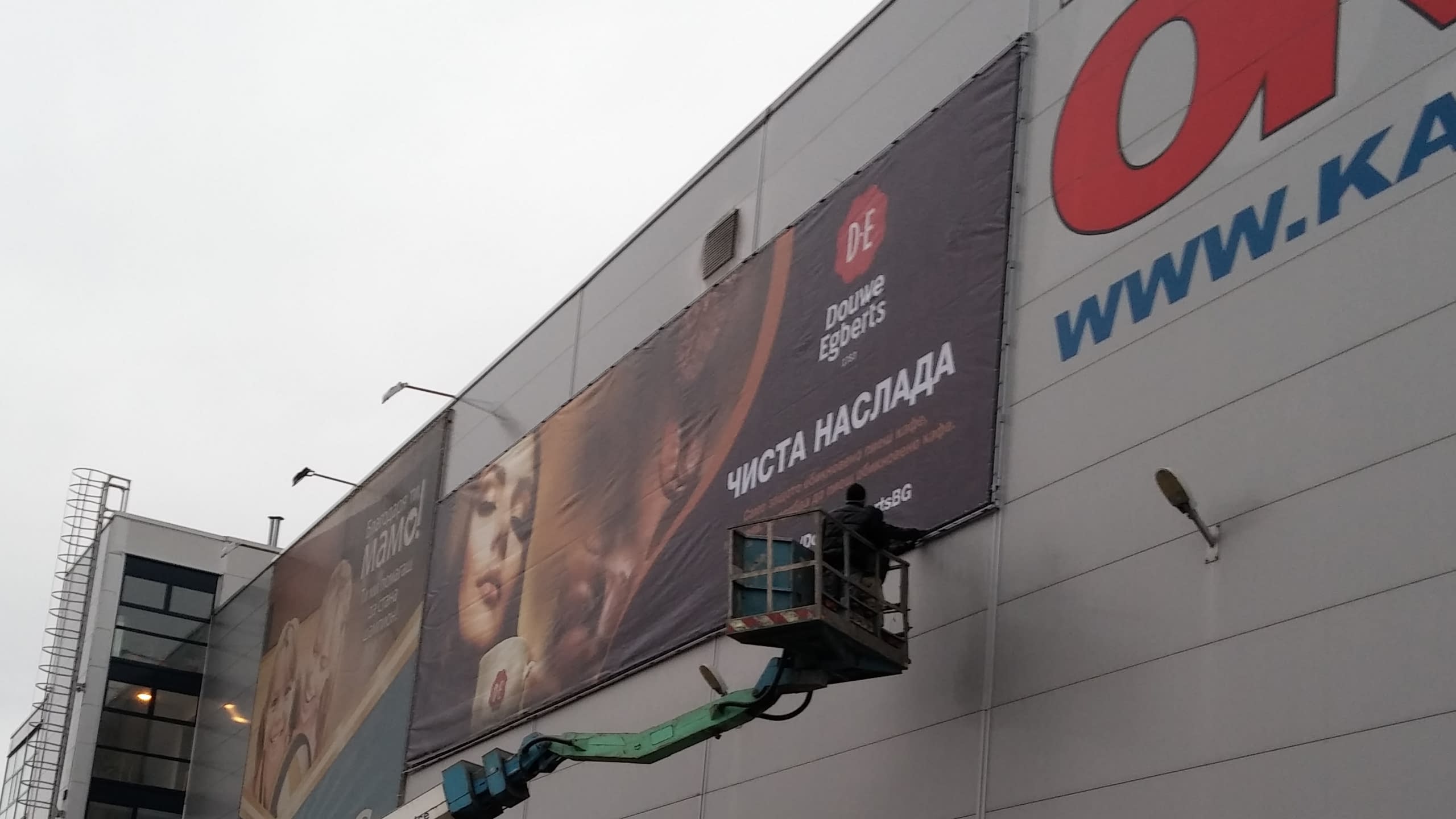Монтаж на голямоформатно рекламно платно на "Douwe Egberts" на фасада на търговски обект. Работник на вишка извършва монтажа.