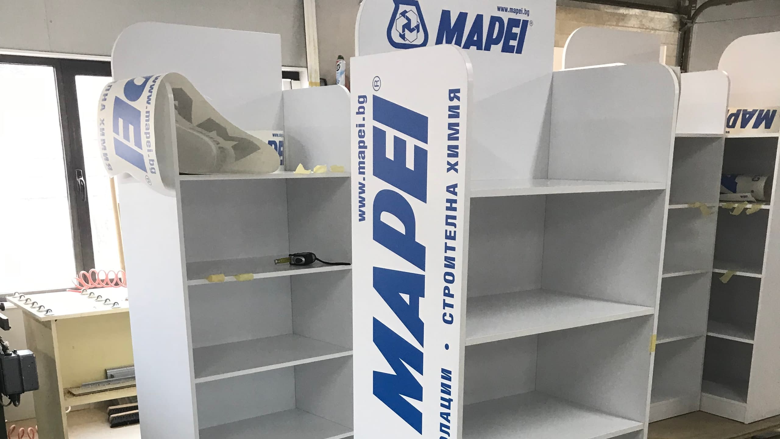 Изработка и брандиране на бели рекламни щендери за строителна химия "MAPEI". Процес на облепване на стелажи с рекламно фолио с логото на фирмата.