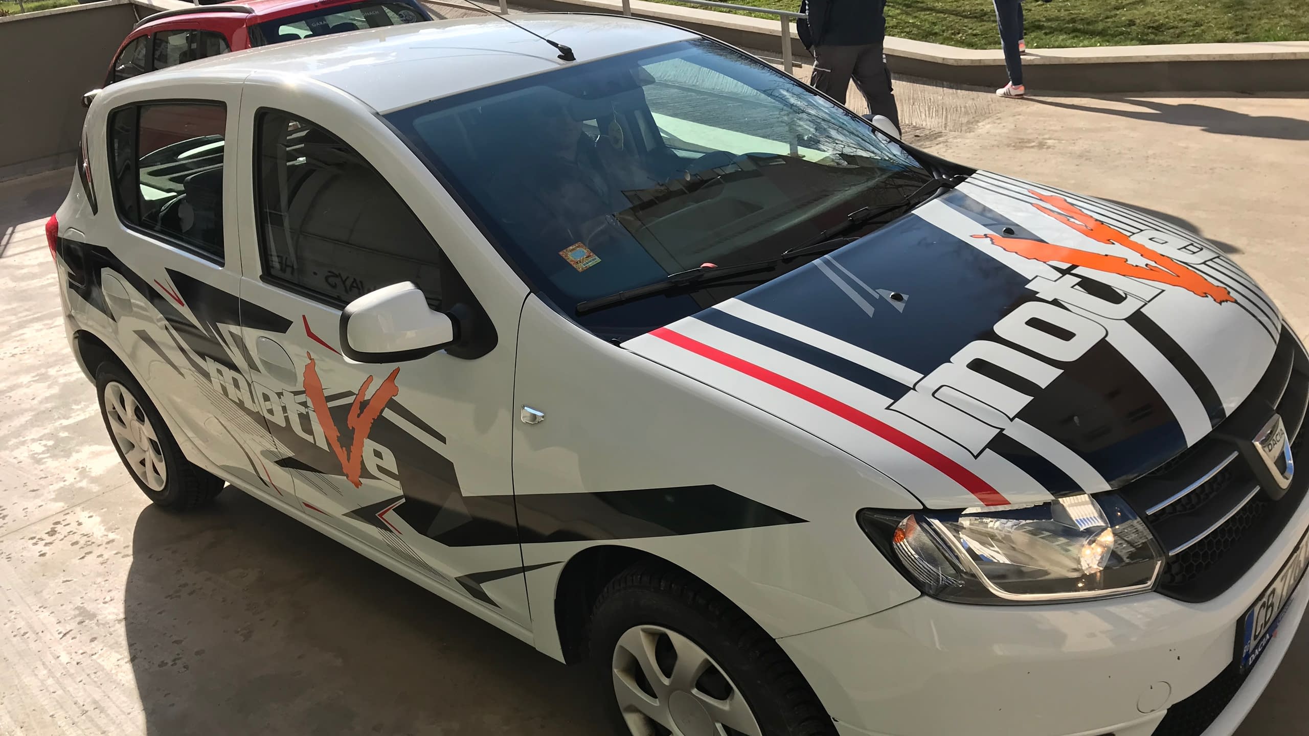 Бял автомобил Dacia с частично брандиране с рекламни стикери. Дизайнът включва динамични графики в черно, оранжево и червено с лога на "Motul" и "mobile v"