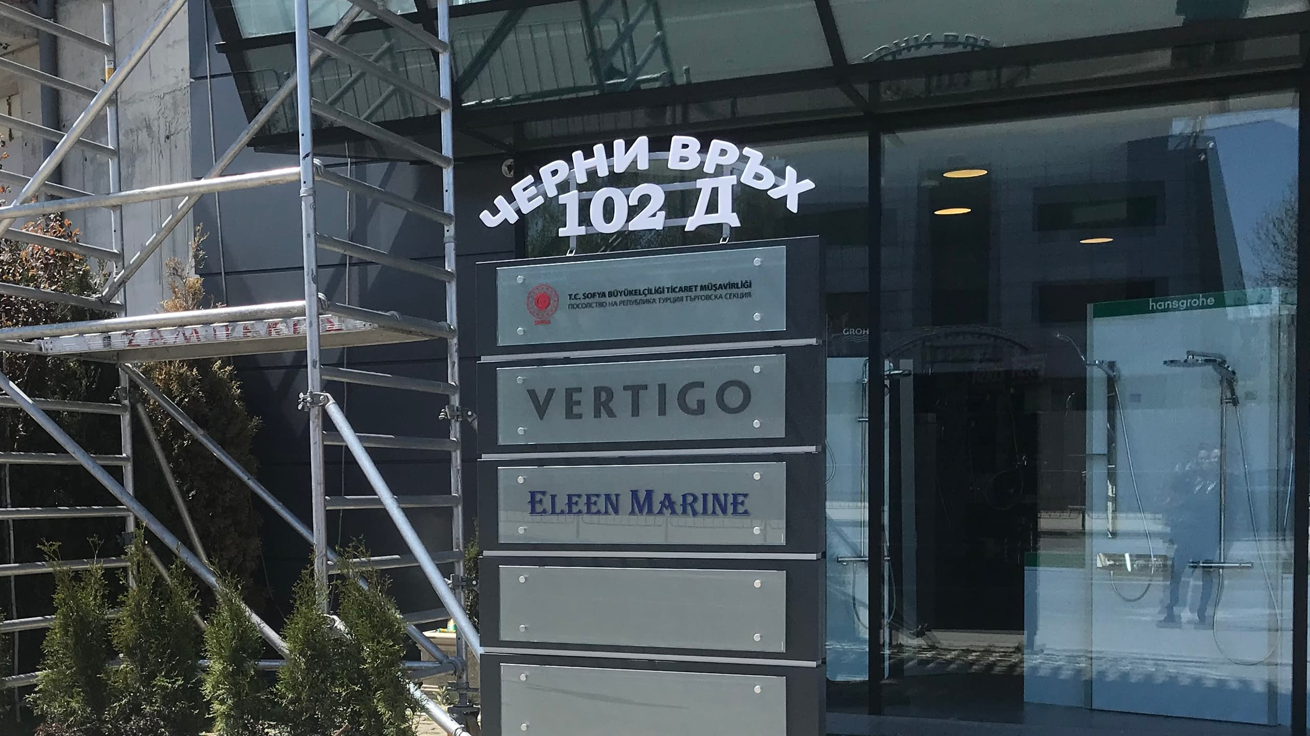 Надписи от бяло рязано фолио на стъклена врата на офис сграда с адрес "Черни връх 102 Д" и указателни табели с имена на фирми.