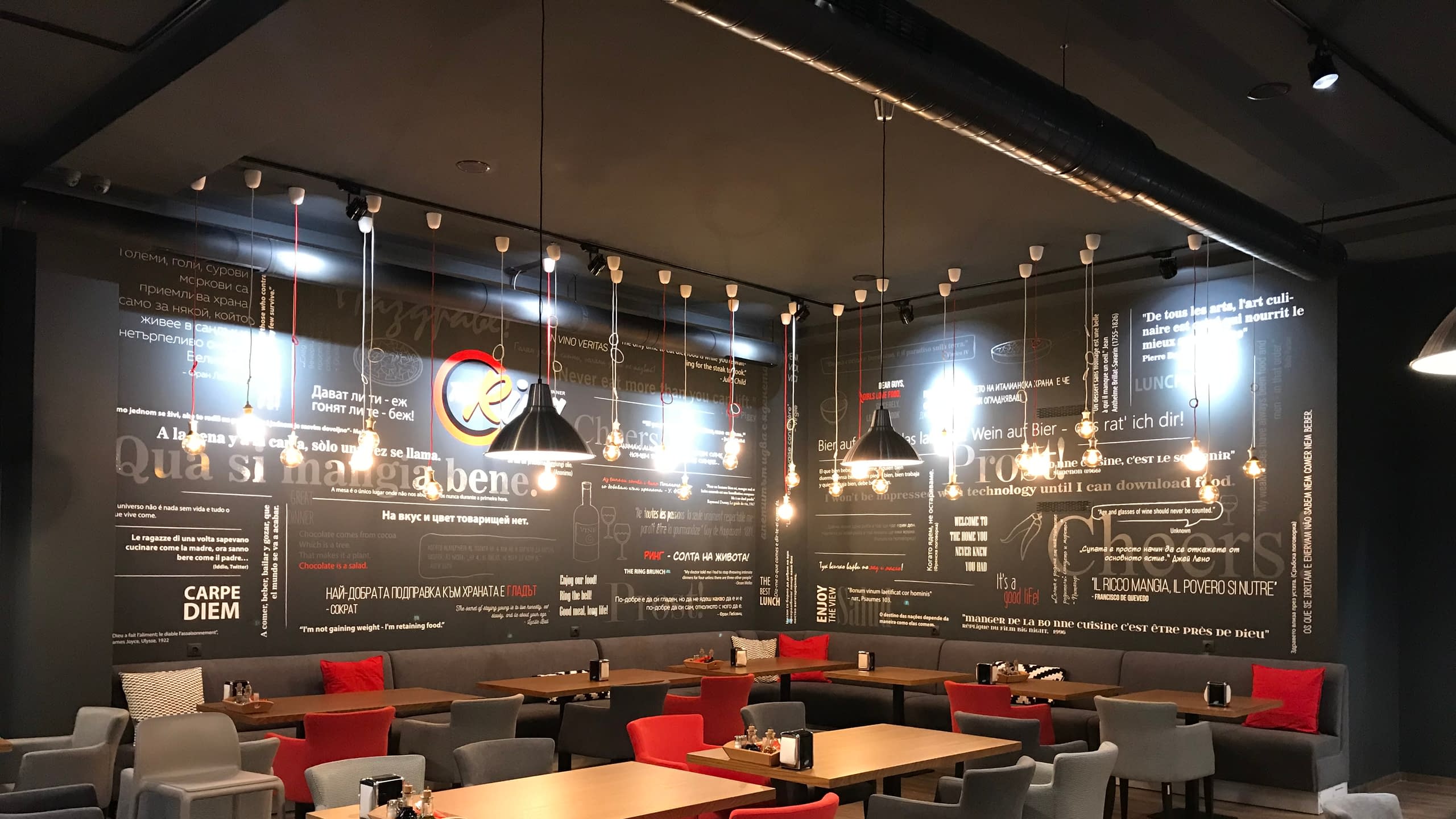 Интериор на модерен ресторант със стена, декорирана с типографски надписи от рязано фолио на различни езици. Декорацията създава артистична атмосфера.