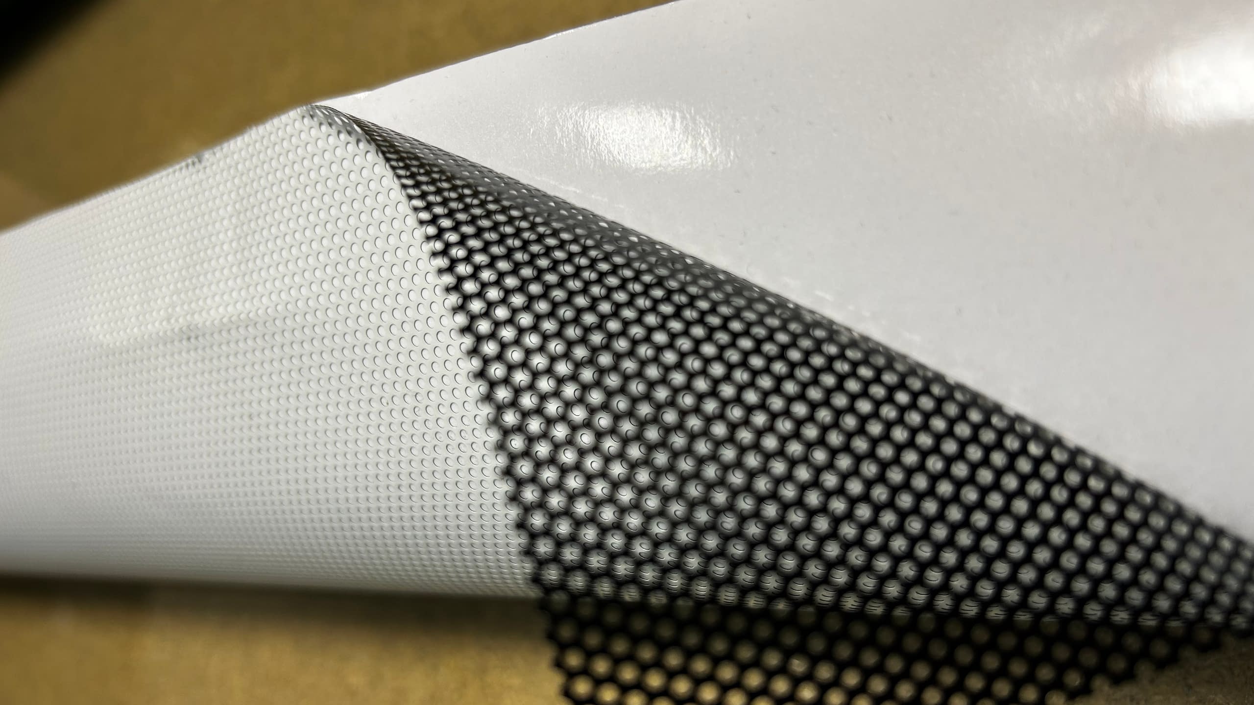 Близък план на перфо фолио (one-way vision) за стъкла, което се отлепя от подложката си. Вижда се перфорираната структура на материала.