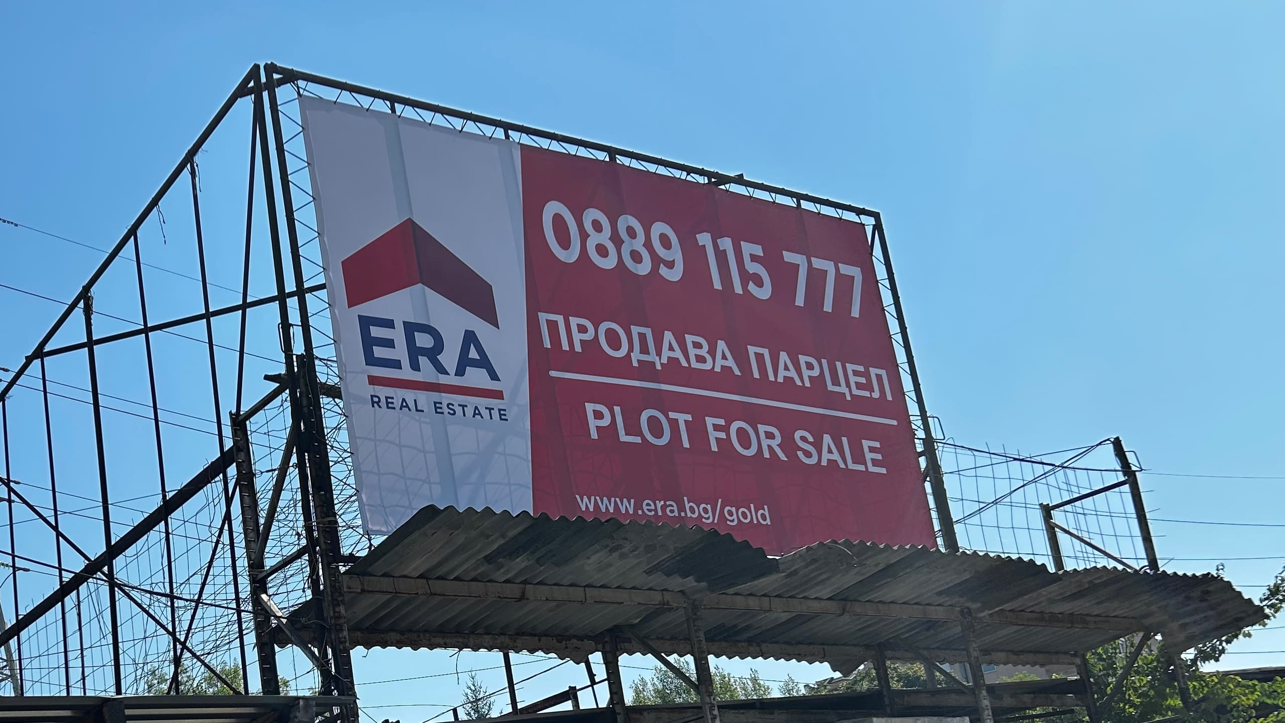 Рекламен билборд на "ERA Real Estate" с обява за продажба на парцел. Банерът съдържа логото на компанията, телефонен номер и уебсайт.