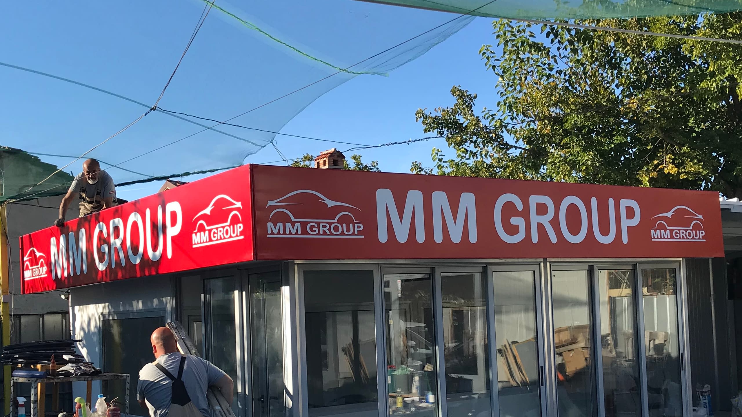 Монтаж на червена светеща рекламна табела с лого на "MM GROUP" върху покрива на търговски обект. Двама работници монтират конструкцията.