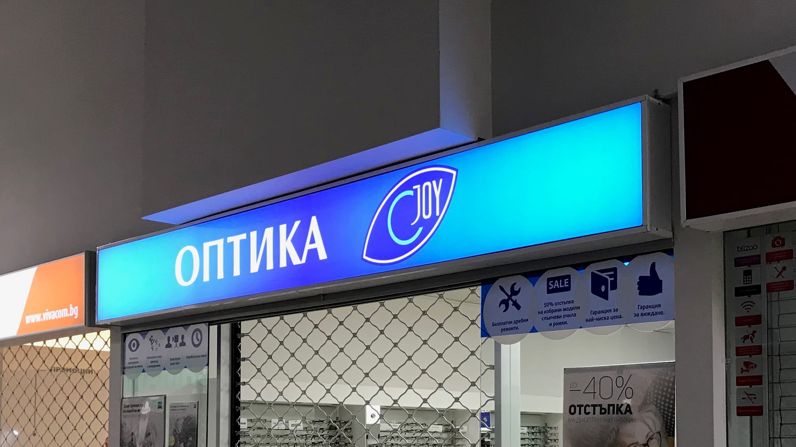 Правоъгълна светеща кутия (лайтбокс) със син фон и бял надпис за "Оптика JOY", монтирана над входа на търговски обект.