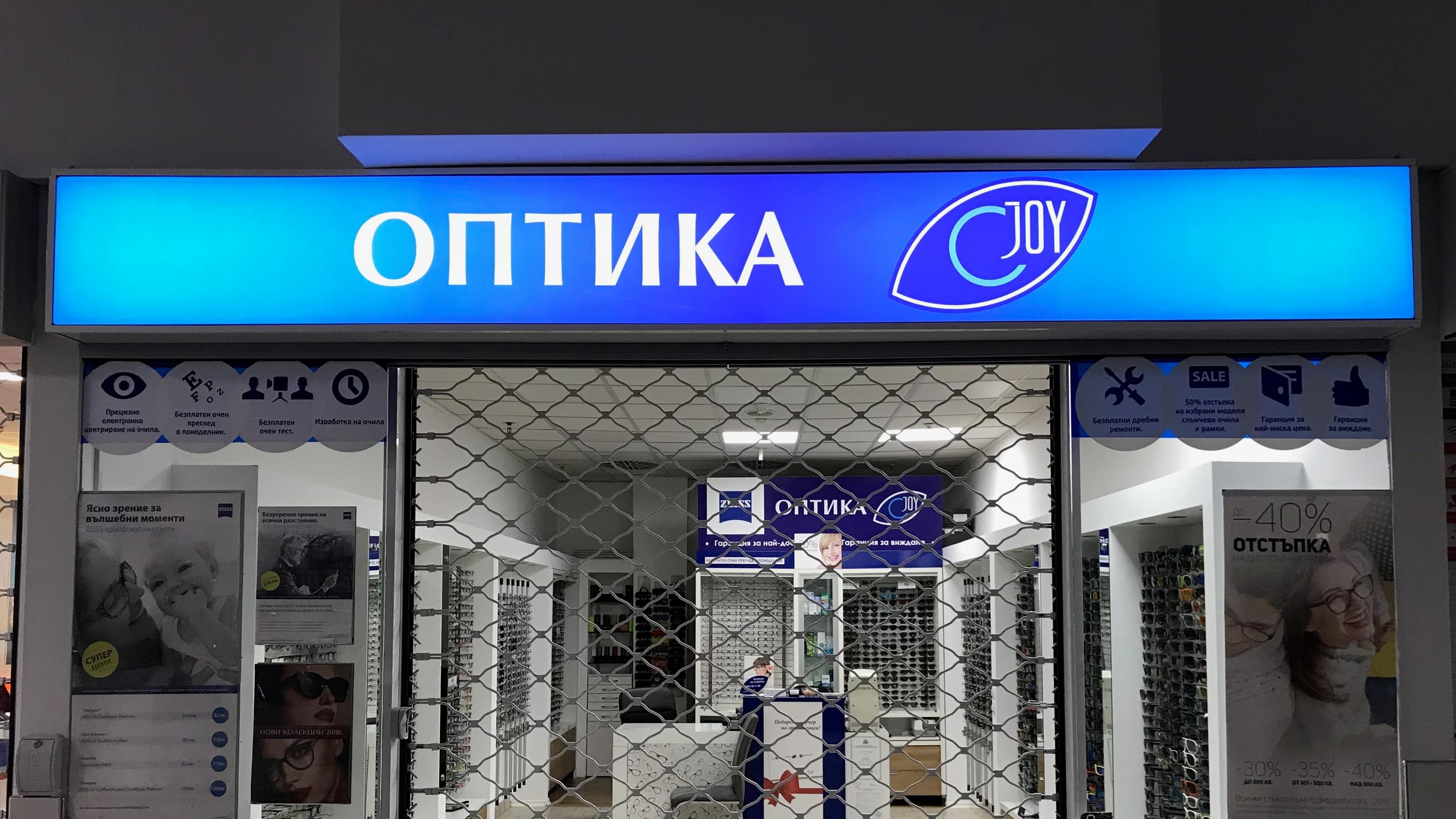 Светеща табела с винил на магазин "Оптика JOY", светеща в яркосиньо пред спуснати охранителни щори.