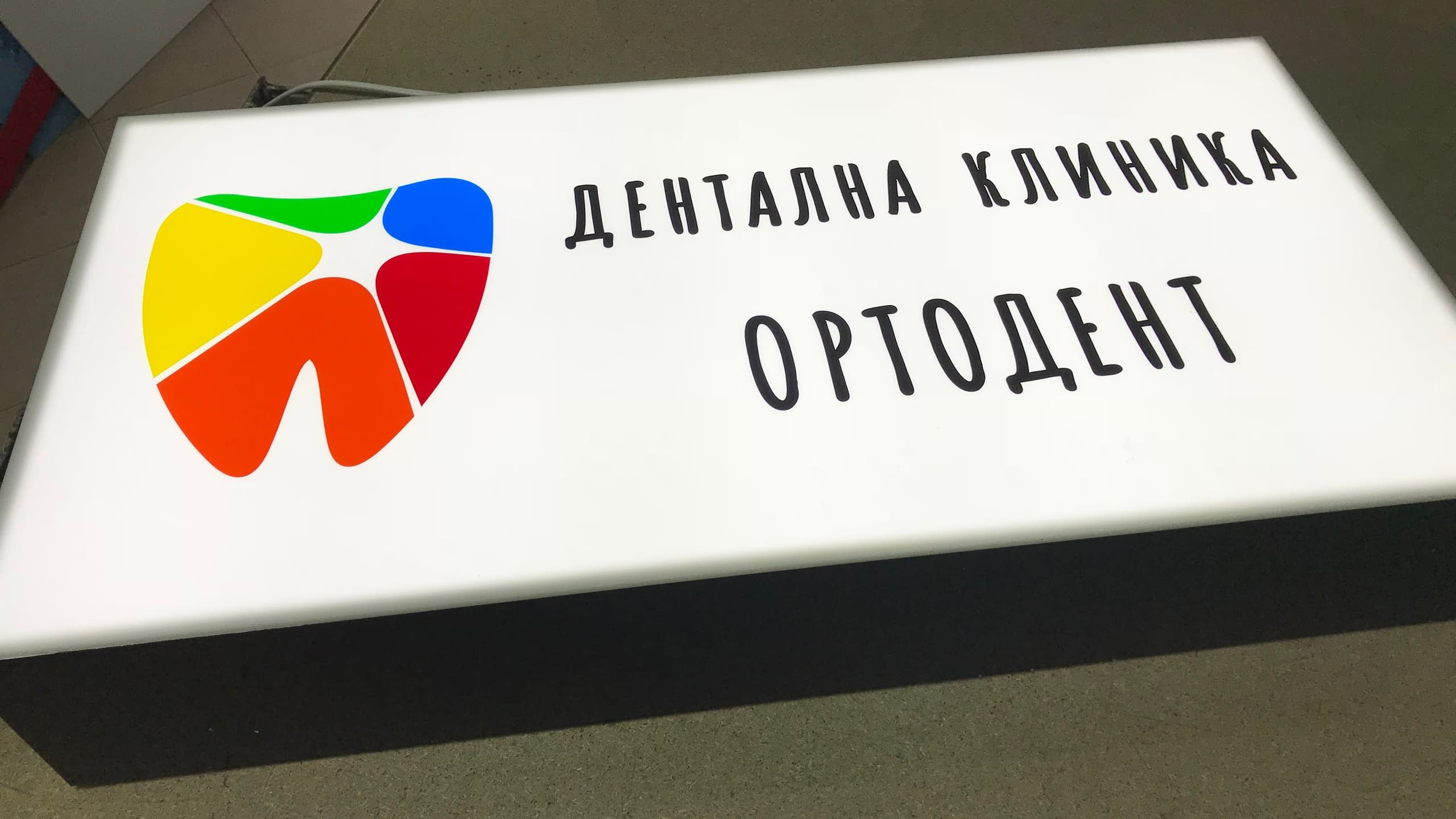 Бяла светеща кутия с равномерно осветление за "Дентална клиника ОРТОДЕНТ", с цветно лого във формата на зъб и черен надпис.