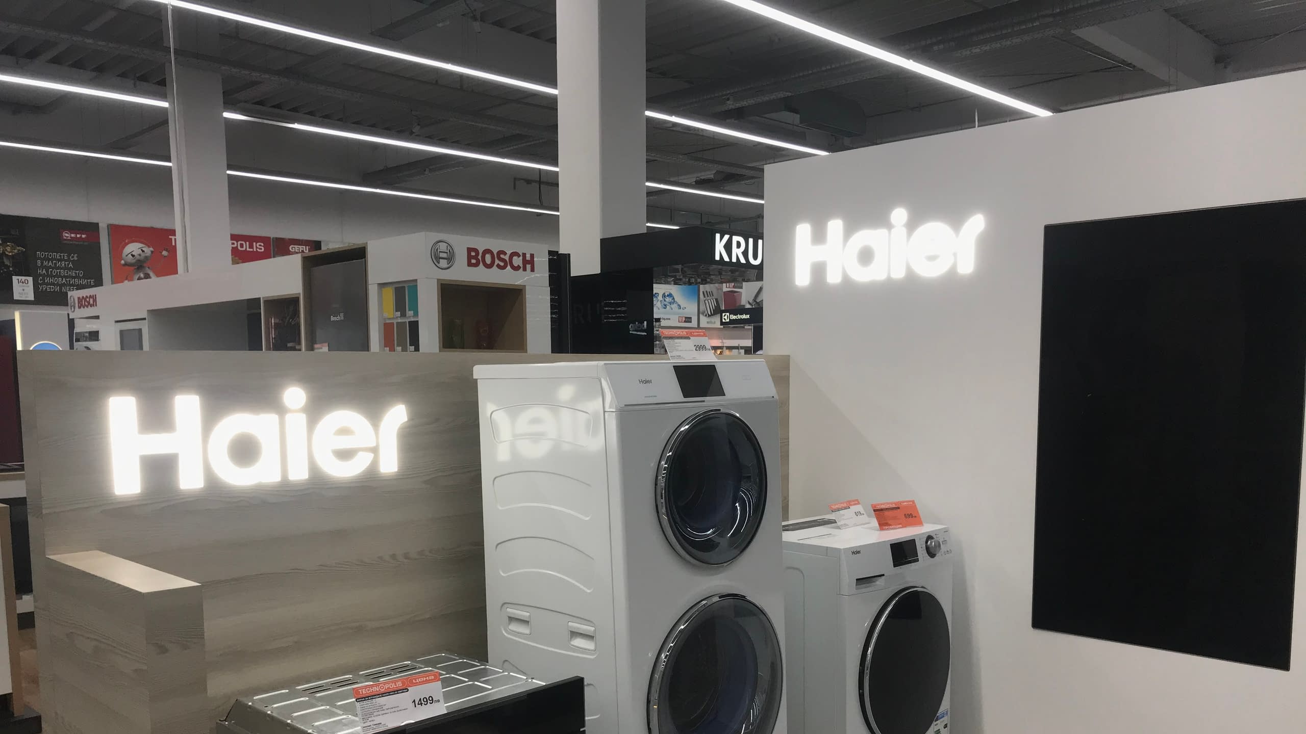 Интериорни светлинни реклами с логото на "Haier" в голям магазин за електроуреди, позиционирани до продуктите на марката.