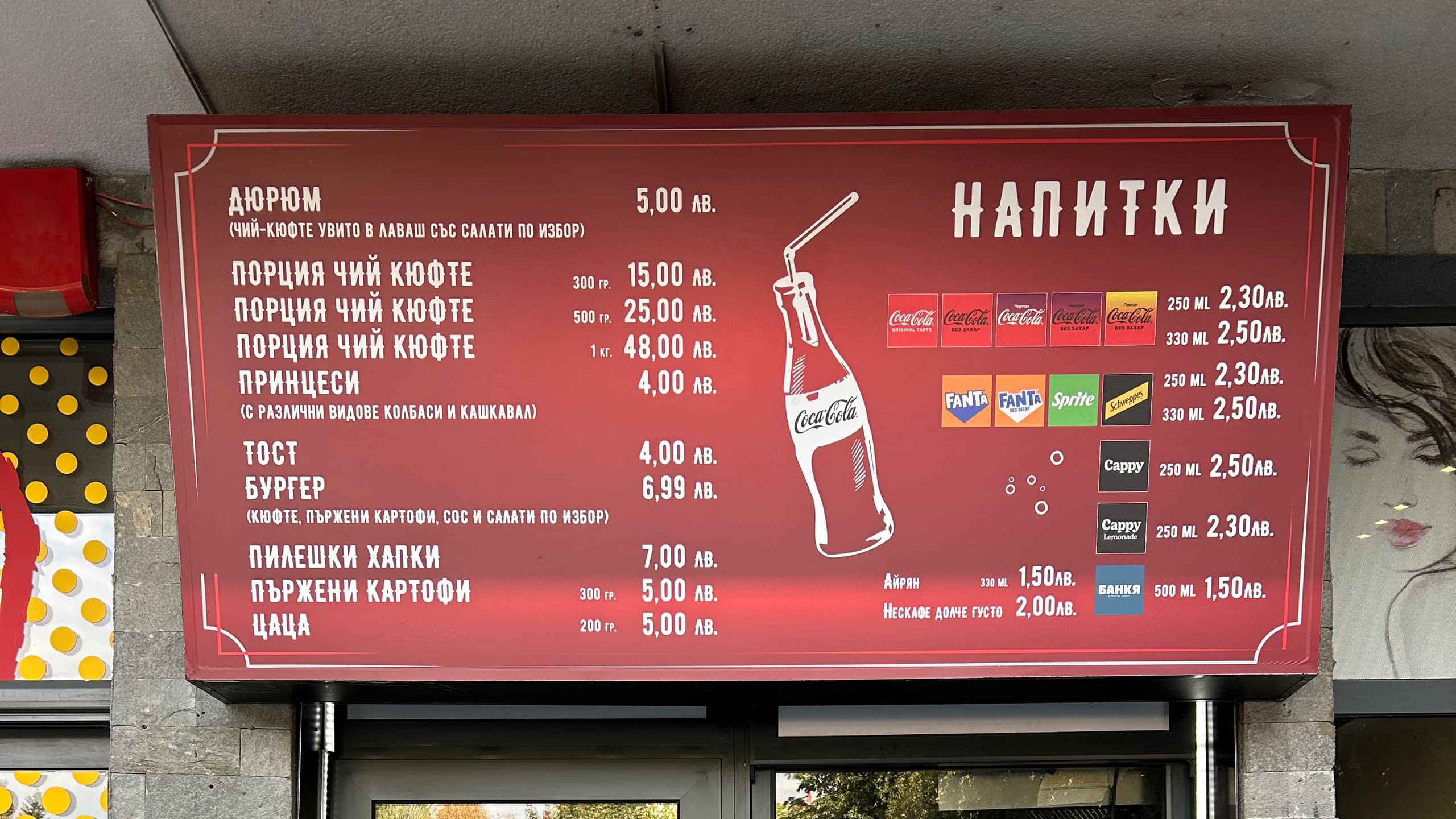 Голяма рекламна табела-меню за заведение за бързо хранене с изписани ястия, напитки и цени, монтирана на открито.