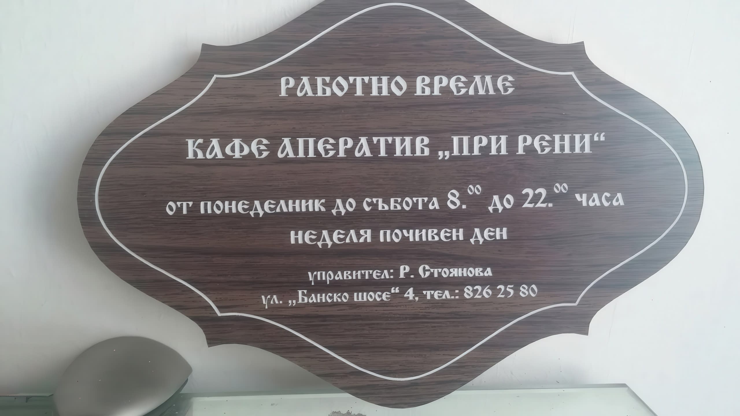 Фигурна гравирана табела с ефект на тъмно дърво, указваща работното време и контактите на "Кафе аператив „При Рени“".