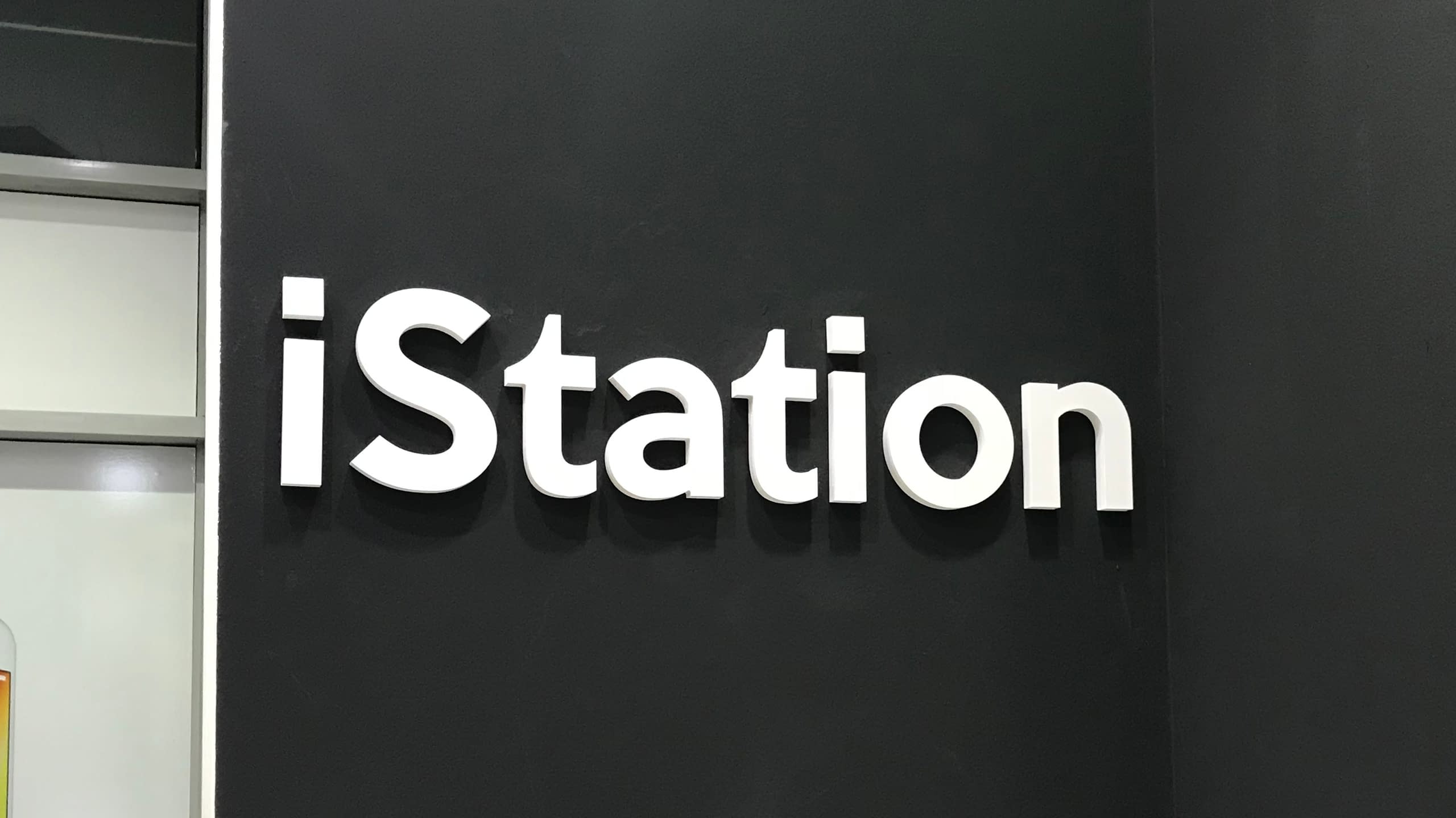 Интериорна табела с бели обемни букви "iStation", монтирани на тъмна стена в магазин или офис.
