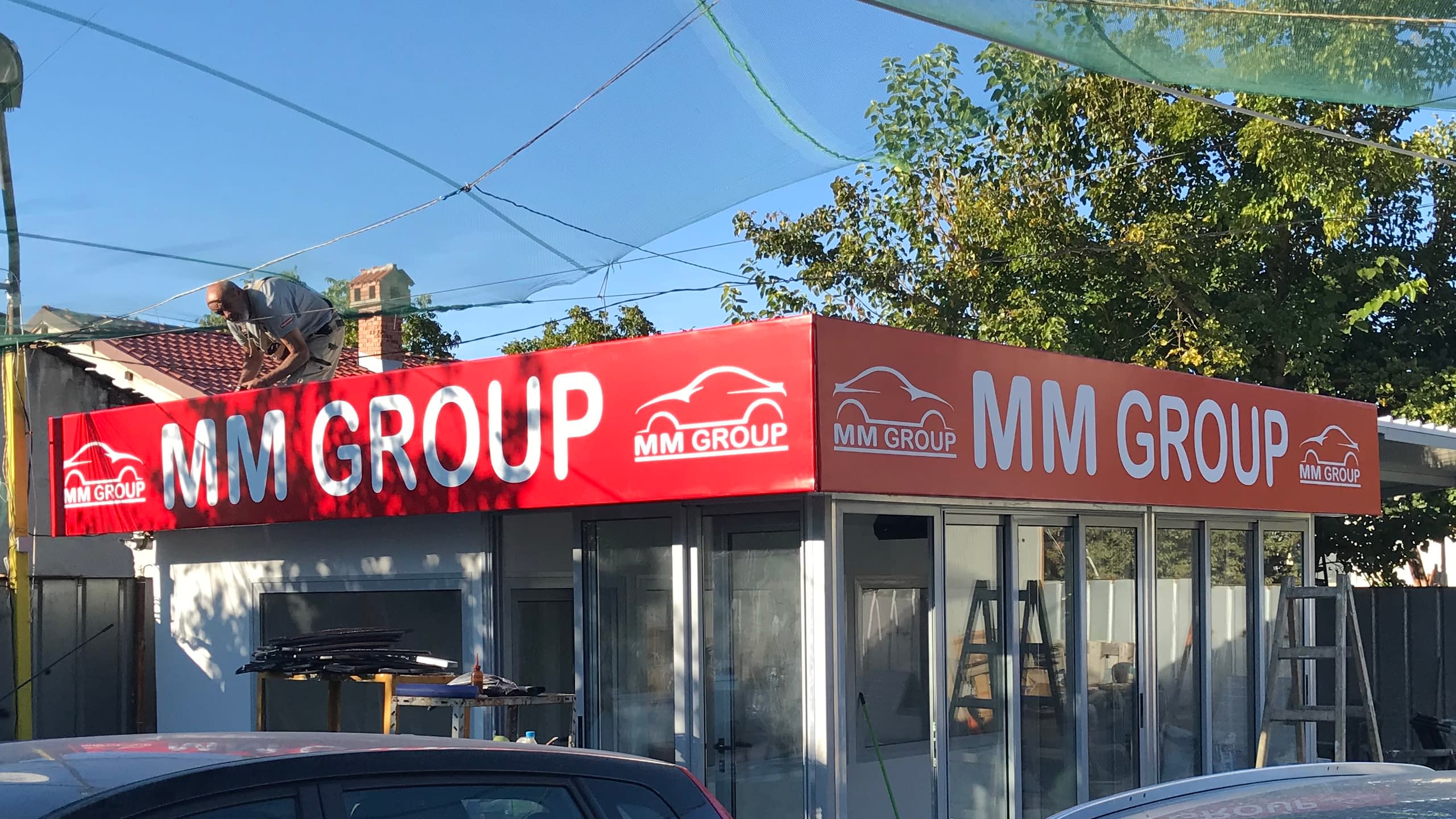 Светеща винилна реклама в червено с логото на "MM Group", обгръщаща фриза на търговски обект по време на монтаж.